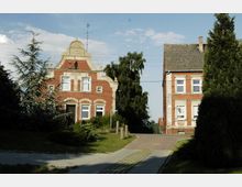 Zwei rot verklinkerte historische Wohngebäude mit weißen Fensterrahmen, eines mit verziertem Giebel, stehen nebeneinander an einer gepflasterten Zufahrtsstraße. Im Vordergrund sind Rasenflächen und Bäume zu sehen, der Himmel ist leicht bewölkt.