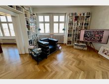 Wohnzimmer mit hellen Holzboden in Fischgrätmuster, ausgestattet mit hohen Bücherregalen, einem schwarzen Ledersessel und einem kleinen Glastisch. Große Fenster lassen viel Tageslicht herein, und ein Flachbildfernseher auf einem Holzstativ steht an der rechten Wand.