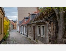 Schmale, gepflasterte Gasse mit alten, eng aneinander stehenden Häusern, teils mit verfallenen Fassaden und roten Ziegeldächern; links gelber blühender Strauch, rechts ein großer Baum. Im Hintergrund sind weitere Gebäude und die Spitze eines Kirchturms sichtbar.