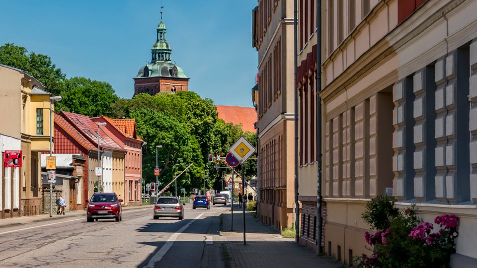 Straßenszene in einer Altstadt mit zweispuriger Fahrbahn, Gehweg und dicht stehenden, teils farbigen Gebäudefassaden mit Satteldächern. Im Hintergrund ragen Bäume und der Turm einer Kirche über die Häuser, entlang der Straße stehen Verkehrsschilder und einige geparkte bzw. fahrende Autos.