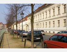 Straßenszene mit beidseitig parkenden Autos, einer hellen historischen Häuserreihe mit rechteckigen Fenstern rechts und einer Baumreihe sowie gepflastertem Gehweg links. Am Horizont ist ein großes Gebäude mit rotem Dach erkennbar.
