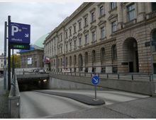Einfahrt zu einer Tiefgarage mit einem Schild, das auf "Parkhaus öffentlich" und "FREI" hinweist, vor einem historischen Gebäude mit sandfarbener Fassade und großen, bogenförmigen Fenstern. Im Hintergrund sind ein weiteres Gebäude mit grünlicher Kuppel und Nationalflaggen sichtbar.