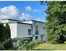 Moderne, rechteckige Doppelhaushälfte mit weißer Fassade und Flachdach, umgeben von Bäumen und Büschen. Im Vordergrund befindet sich ein Gartenbereich mit grünem Rasen und sichtbarem Zaun.