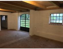 Innenraum eines renovierten historischen Gebäudes mit weiß verputzten Wänden, einer großen Glastür mit schwarzen Sprossen, Ziegelboden und Holzbalken an der Decke. Durch die Fenster ist eine grüne Wiese sichtbar.