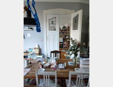 A cozy interior showing a cluttered wooden dining table with chairs, surrounded by shelves and cabinets containing books and small items. Double white doors with decorative stained glass panels lead to a room with more bookshelves and toys, highlighting a lived-in and creative atmosphere.