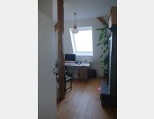 Ein kleiner, heller Dachraum mit sichtbaren Holzbalken, einer schräge Fensterfront, Holzboden und einem Schreibtisch, auf dem Arbeitsmaterialien liegen. Im Raum steht eine große Pflanze, und ein schwarzer Schrank ist sichtbar.