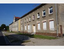 Straßenansicht eines Reihenhauses mit verputzter Fassade, sichtbaren Schäden und geschlossenen Rollläden an den Fenstern. Der Gehsteig und eine unbefestigte Fläche befinden sich vor dem Gebäude, das in einer ruhigen Umgebung mit vereinzelten Bäumen liegt.