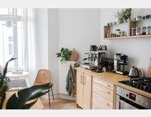 Helle Küche mit Holzarbeitsflächen, Kaffeemaschinen und Utensilien auf der rechten Seite, einem großen Fenster mit weißen Vorhängen links und einem Stuhl aus geflochtenem Material vor einem Heizkörper mit Pflanzen und Geschirrtuch.