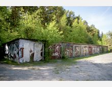 Reihe alter Garagen mit Wellblechdächern, deren Türen mit Graffiti bemalt sind, umgeben von dichtem Wald mit grünen Bäumen; im Vordergrund eine unbefestigte Fläche.