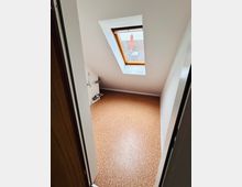 Kleiner Dachgeschossraum mit schrägen weißen Wänden und Korkboden, gesehen durch eine Türöffnung. Ein Dachfenster mit Holzrahmen lässt Tageslicht herein und bietet Blick auf ein Ziegeldach; an der linken Wand befindet sich ein Heizkörper.