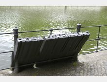 Schwarzes Metalldenkmal mit der Aufschrift „Rosa Luxemburg“ am Ufer eines Gewässers, eingefasst von einem Geländer aus Metallstreben. Der Hintergrund zeigt grünes, leicht bewegtes Wasser.