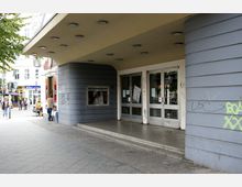 Eingangsbereich eines Gebäudes mit grauer, geschwungener Fassade und einem breiten Vordach. Doppeltüren aus Glas und ein Schaukasten sind sichtbar, teilweise mit Graffiti versehen; im Hintergrund eine belebte Straßenszene.