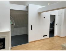 Weiß gestrichener Innenraum mit Holzboden, einem eingebauten Kamin an der linken Seite und zwei offenen Türbereichen; der Bereich links zeigt weiße Fliesenwände und ein Rundbogenfenster, während rechts ein Badezimmer mit Schrägdecke und Fliesen sichtbar ist.