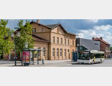 Ziegelgebäude eines historischen Bahnhofs mit großen Fenstern, daneben ein moderner Anbau mit der Aufschrift „Museum“. Im Vordergrund ein überdachter Buswartebereich, eine Bushaltestelle und ein ankommender Linienbus vor gepflastertem Platz.