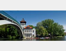 Ein historisches Gebäude mit roten Dächern und Fachwerkelementen steht am Ufer eines Flusses, umgeben von Bäumen. Im Vordergrund spannt sich eine weiße Bogenbrücke, und am Wasser liegen bunte Kanus.