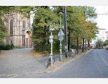 Straßenansicht mit gepflastertem Gehweg, einer historischen Kirche aus Backstein links mit großem Eingangsportal, umgeben von Bäumen, und einer Straßenecke mit parkenden Fahrrädern sowie Straßenbahnschienen rechts. Im Hintergrund sind Wohnhäuser und Geschäfte zu sehen.