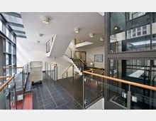 Moderne Treppenhausansicht mit Glasgeländern, schwarzen Fliesenböden und einem großen Fensterbereich auf der linken Seite, der viel Tageslicht einlässt. Mehrere Treppenabschnitte mit Holzhandläufen führen in verschiedene Etagen, und im Vordergrund steht ein Metallwagen.