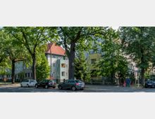 Straßenszene mit mehrstöckigen Wohnhäusern im Hintergrund, teilweise in Pastellfarben gestrichen und mit roten Ziegeldächern. Große Bäume säumen die Straße, entlang derer Autos geparkt sind, und eine Person mit einem Kinderwagen geht auf dem Gehweg.