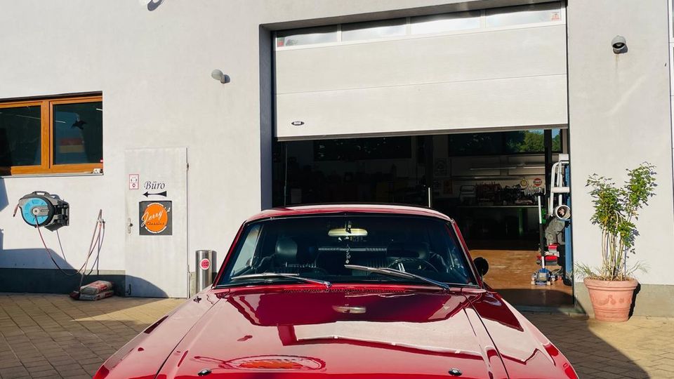 Außenansicht einer Werkstatt- oder Garagenhalle mit grauer Fassade und roter Dachkante, großem Rolltor und Firmenlogo über dem Eingang. Vor dem geöffneten Tor steht auf einem gepflasterten Hof ein roter Oldtimer, daneben sind ein Fenster, ein Seiteneingang und ein Blumentopf zu sehen.