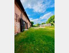 Eine weitläufige Wiese mit einer alten Backsteinmauer auf der linken Seite, die Teil eines Gebäudes mit großen Holztoren ist. Im Hintergrund sind ein Gewächshaus und Bäume unter einem teils bewölkten blauen Himmel zu sehen.