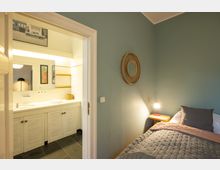 Schlafzimmer mit mintgrünen Wänden, einem Bett mit grauer Decke und Nachttisch mit Lampe. Im Hintergrund ein offenes Badezimmer mit weißem Waschtisch, Spiegelschrank und gelben Regalen.