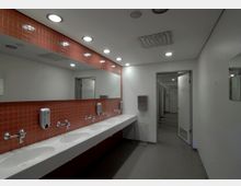 Öffentliche Badezimmeranlage mit einer langen weißen Waschrinne, daran befestigte Armaturen und Seifenspender vor einer roten Kachelwand mit großem Wandspiegel. Rechts im Bild sind Türen mehrerer Toilettenkabinen zu sehen.