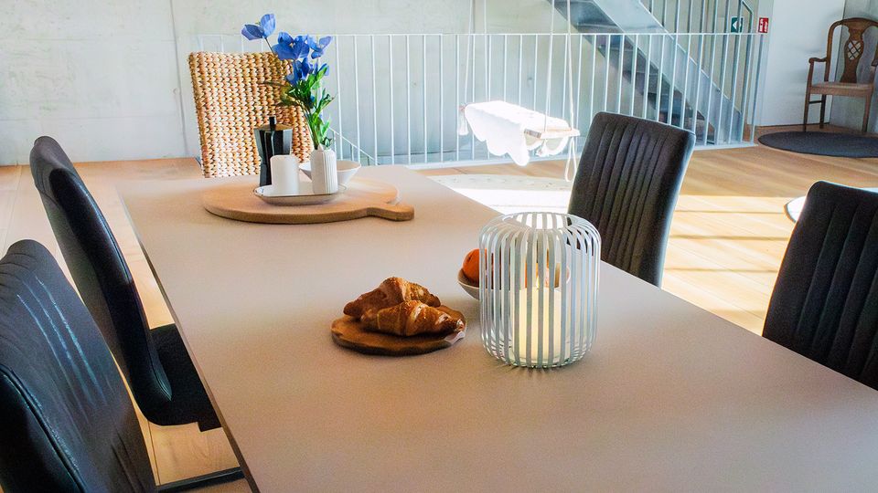 Heller, moderner Essbereich mit großem rechteckigem Tisch und dunklen Polsterstühlen, dekoriert mit Kerzen, Blumen und Frühstücksgebäck. Im Hintergrund sind ein offenes Treppengeländer, helle Holzböden und ein einzelner Stuhl an der Wand zu sehen.