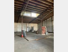 Leerer Innenraum eines alten Industriegebäudes mit sichtbaren Holzbalken an der Decke, Metallstützen, rauen Wänden aus Mauerwerk und einigen baulichen Elementen wie einer roten Tür und Bauzubehör. Licht fällt durch Fenster im oberen Bereich der Wand ein.