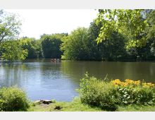 Ein ruhiger See inmitten eines Parks, umgeben von dichtem, grünem Laub und blühenden Pflanzen am Ufer; im Hintergrund paddeln Menschen in einem roten Boot.