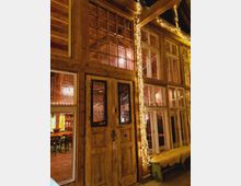 Holzfassade eines Gebäudes mit großen Fenstern und einer verzierten Doppeltür. Die Fassade ist mit Lichterketten dekoriert, und im Inneren sind Tische und Stühle sowie warmes, gelbliches Licht sichtbar.