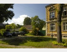 Wohngebiet mit einer gelben Klinkerfassade eines historischen Gebäudes rechts, mehreren geparkten Autos auf einem unbefestigten Parkplatz links und grüner Vegetation im Vorder- und Hintergrund. Stromleitungen verlaufen über die Szene, und ein Baum sowie ein Laternenpfahl stehen nahe dem Gebäude.