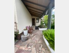 Eingangsbereich eines Hauses mit überdachtem Steinboden. Links stehen Holzmöbel mit gepolsterten Kissen, rechts befinden sich Pflanzen, darunter Farn in einem weißen Blumenkasten. Im Hintergrund ist eine Holztür und ein kleines Fenster zu sehen.