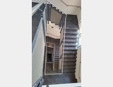 Blick von oben in ein modernes Treppenhaus mit mehreren Etagen, dunkel gefliesten Treppenstufen und Podesten. Metallgeländer mit Füllpaneelen rahmen den Treppenschacht, seitlich fällt Licht durch ein hohes Fenster.