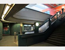 U-Bahnhof-Interieur mit einer Rolltreppe und einer Treppe, flankiert von grünen Fliesenwänden und beleuchteten runden Lampen. Im Hintergrund sind Schilder, Graffiti und ein Geschäft mit großen Fenstern zu sehen.