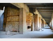 Ehemaliger Industriehallenbereich mit gemauerten Stein- und Ziegelwänden, sichtbaren Rundöfen und massiven Betonbalken, flankiert von hohen, rechteckigen Öffnungen auf einer Seite. Der Boden ist aus Beton, und die Struktur wirkt historisch und rustikal.