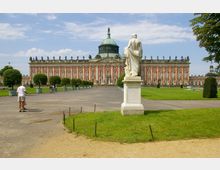 Ansicht des Neuen Palais im Park Sanssouci, Potsdam, mit seiner barocken Fassade in Rot- und Beigetönen, einer zentralen Kuppel und zahlreichen Statuen auf dem Dach; im Vordergrund eine weiße Statue und eine weitläufige, gepflegte Gartenanlage mit gepflasterten Wegen.