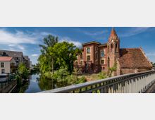 Historisches Gebäudeensemble mit einer Backsteinkirche und einem größeren, mehrstöckigen Backsteinbau neben einem Fluss, umgeben von Bäumen und modernen Wohnhäusern im Hintergrund, aufgenommen von einer Brücke aus.