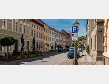Straßenzug in einer historischen Altstadt mit mehrgeschossigen, pastellfarbenen Fassaden und hohen Fenstern, teils mit Caféfront im Erdgeschoss. Am rechten Straßenrand stehen parkende Autos, ein Laternenmast mit Parkhinweisen sowie Gehwege mit Kopfsteinpflaster und kleinen Straßenbäumen.