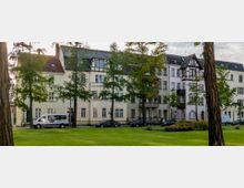 Wohngebäude im historischen Stil mit Fachwerkelementen, großen Fenstern und hellen Fassaden, umgeben von Bäumen und einer gepflegten Grünfläche im Vordergrund; parkende Autos stehen entlang der Straße.