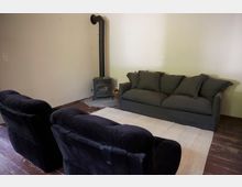 Wohnzimmer mit schlichtem Interieur, bestehend aus einer grauen Couch, zwei schwarzen Sesseln, einem kleinen Holzofen in der Ecke und einem hellen Teppich auf dem dunklen Holzfußboden.