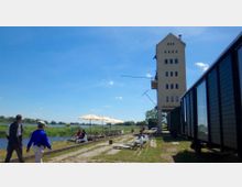 Ein altes Industriegebäude mit schmalem Turm und mehreren Fenstern steht in einer offenen Landschaft am Ufer eines Flusses. Im Vordergrund befinden sich Sitzgelegenheiten mit Sonnenschirmen, ein Weg aus Kopfsteinpflaster und grüne Wiesen; rechts sind dunkle, fensterreiche Waggons zu sehen.
