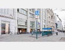 Außenansicht einer städtischen Einkaufsstraße mit dem Eingang zur U-Bahn-Station „Kurfürstendamm“ (U1, U9) im Vordergrund. Umgeben von modernen Gebäuden mit großen Fensterfronten und einem historischen Gebäude mit verzierten Fassaden, sind auch Passanten, Geschäftseingänge und abgestellte Fahrräder zu sehen.