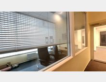 Blick durch ein großes Fenster mit Jalousien in ein Büro mit zwei schwarzen Bürostühlen, einem Schreibtisch und Regalen. Der Raum hat gelbe Wände, Teppichboden und wird von einer anderen Tür im Flur flankiert.