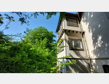 Seitenansicht eines mehrgeschossigen Wohnhauses mit weißer Holzfassade und braunen Fensterrahmen, umgeben von dichtem Grün aus Bäumen und Sträuchern unter einem klaren blauen Himmel.