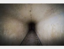 Ein schmaler, langer Tunnel mit abgenutzten, beige-grauen Wänden und einem leicht gewölbten Deckenverlauf. Der Betonboden ist uneben, und am Ende des Tunnels ist ein dunkler Durchgang zu erkennen.