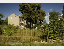Ein Wohnhaus mit heller Fassade und mehreren Fenstern steht hinter einem verwilderten Vorgarten mit hohem Gras und Bäumen. Im Vordergrund ist ein schmiedeeiserner Zaun mit einem Tor zu sehen.