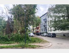 Straßenszene in einem Wohngebiet mit mehrstöckigen, farbig gestrichenen Altbauten mit Balkonen auf der rechten Seite. Vor den Häusern stehen geparkte Autos, und links befindet sich eine grüne Fläche mit Bäumen und Sträuchern sowie einer Straßenlaterne.