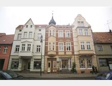 Straßenansicht einer historischen Häuserreihe mit drei mehrstöckigen Gebäuden, die ornamentale und klassische Fassadendetails aufweisen. Das mittlere Gebäude trägt ein Ladenschild mit der Aufschrift "Bernhardt", und im Vordergrund ist eine gepflasterte Straße mit geparkten Autos sichtbar.