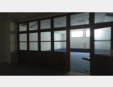 Ein leerer Raum mit weiß gestrichenen Ziegelwänden und einem blauen Teppichboden, getrennt durch eine große Holztür mit mehreren Glasfenstern. Im Hintergrund sind Heizkörper und schmale Fenster zu sehen, die Tageslicht einlassen.