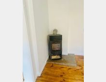 Schwarzer Kaminofen in einer Zimmerecke, auf einer rechteckigen Betonplatte vor weißer Wand. Der Raum hat einen Holzdielenboden und ein kurzes Abzugsrohr führt nach oben in die Wand.